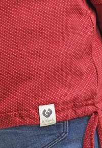 Röd texturerad tyg med små vita prickar, med en knuten nederkant och en etikett som säger "I jorden vi litar" nära sömmen.