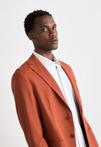 Orange texturerad kavaj med slagkragar och två knappar, tillsammans med en vit skjorta med krage. Minimalistisk design och strukturerad passform.