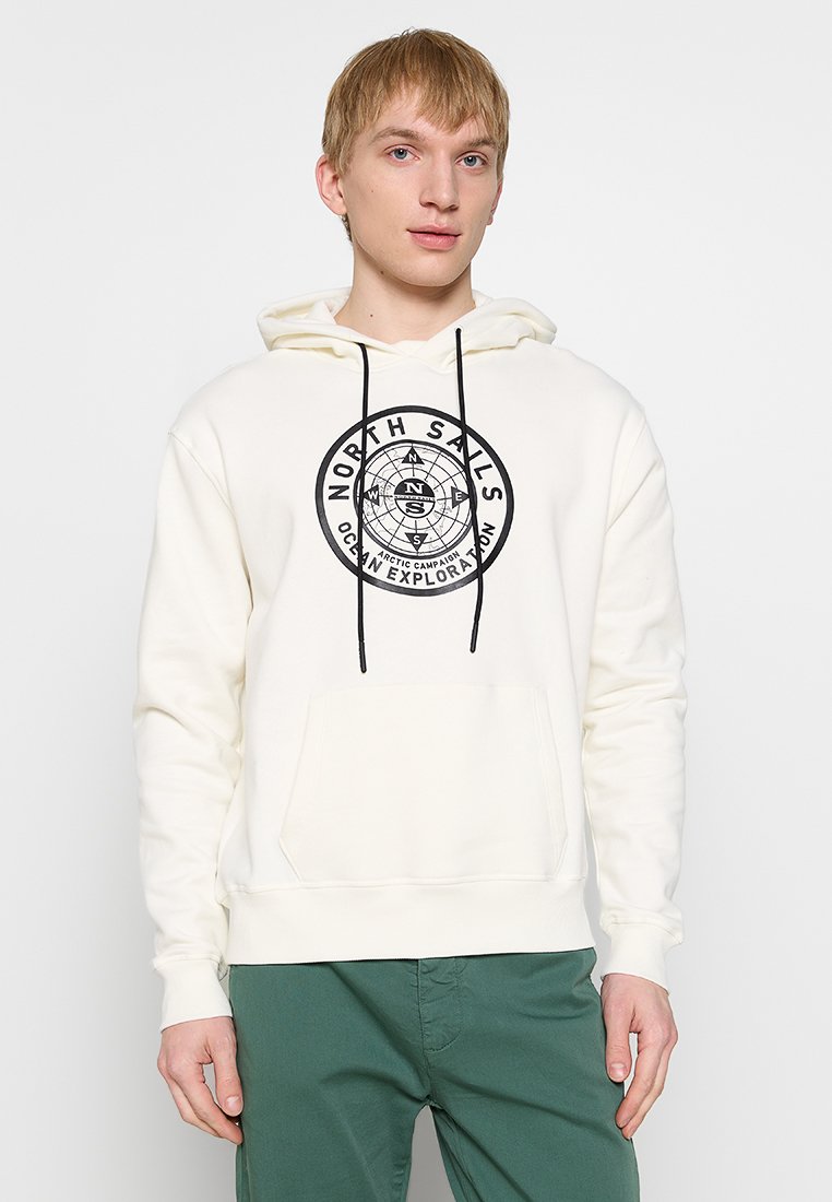 Joven con sudadera blanca con logotipo negro de brújula y texto "North Sails Ocean Exploration" y pantalones verdes, de pie contra un fondo sencillo.