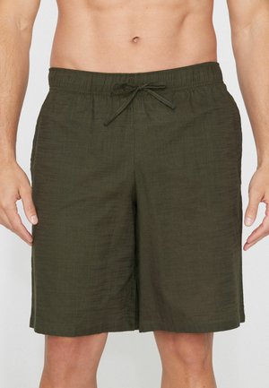 Homme portant un short vert foncé à cordon de serrage, debout, mains détendues sur les côtés, torse et parties supérieures des jambes visibles.