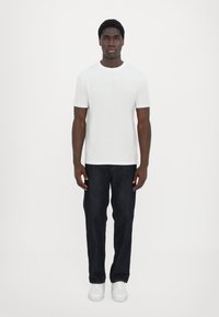 Hvit kortermet t-skjorte med preget logo, kombinert med mørk denimjeans og hvite sneakers. Modellen står vendt mot kameraet foran en ensfarget bakgrunn.