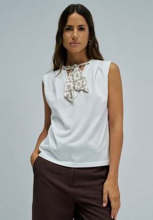 Femme aux longs cheveux foncés portant un haut blanc sans manches avec une écharpe à motifs nouée autour du cou et un pantalon marron foncé sur fond gris.