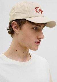 Beige bomulds-kasket med rødt broderet "Origin" tekst, med en buet skygge. Modellen bærer en lys cremefarvet t-shirt, der viser en naturlig hudtone.