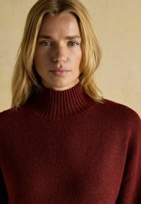 Burgunderfarbener gerippter Rollkragenpullover aus strukturiertem Strick, mit hohem Kragen und langen Ärmeln, vor einem neutralen Hintergrund präsentiert.