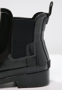 Botte de pluie noire brillante à la cheville avec panneau latéral élastique et languette de traction sur fond blanc.