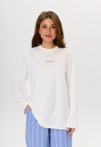 Weißes Sweatshirt mit langen Ärmeln, klassischem Rundhalsausschnitt und pinkem Schriftzug "MIRONS". Kombiniert mit hellblauen gestreiften Hosen.