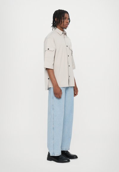 Chemise courte à manches courtes beige clair avec un front à boutons, deux poches poitrine, associée à un jean bleu clair ample et des chaussures noires.