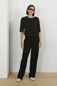 Top negru tricotat cu mâneci scurte, asociat cu pantaloni cu dungi albe și negre. Modelul poartă ochelari de soare rotunzi cu nuanțe de violet și pantofi albi.
