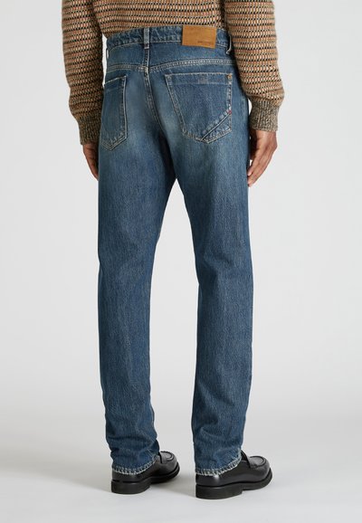 Hombre que lleva vaqueros de mezclilla azules y zapatos de cuero negros, visto desde atrás sobre un fondo blanco liso.