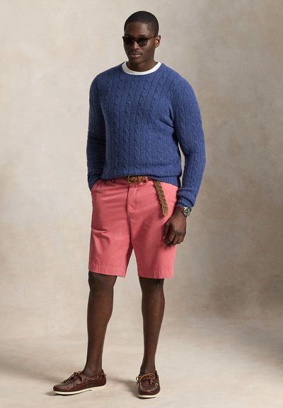 Mann mit Sonnenbrille, blauem Zopfpullover, pinken Shorts mit geflochtenem Gürtel und braunen Leder-Bootsschuhen, stehend vor neutralem Hintergrund.