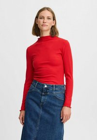 Rotes geripptes Langarmoberteil mit einem hohen Kragen, kombiniert mit einem dunklen Denim-A-Linien-Rock mit einem geknöpften Bündchen und Rüschen an den Ärmelenden.