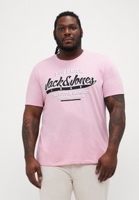 Rozā kokvilnas T-krekls ar melnu uzrakstu "Jack & Jones" un papildus tekstu. Īsas piedurknes un apaļa kakla izgriezuma forma. Vienkāršs dizains.