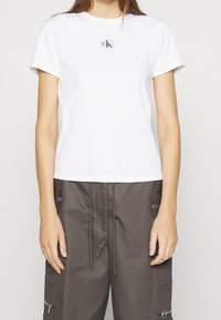 Bílý bavlněný tričko s kulatým výstřihem a krátkými rukávy, s malou černou nášivkou na přední straně. Doplněné hnědými cargo kalhotami.