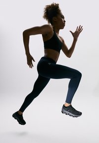 Svart atletisk sportbh och åtsittande marinblå leggings bärs av en person som springer i svarta texturerade skor mot en ljus bakgrund.