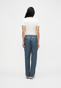 Γυναίκα με μακριά μαύρα μαλλιά, που φοράει εφαρμοστό λευκό τοπ με κοντά μανίκια, φαρδιά μπλε παντελόνια με τσέπες και λευκά αθλητικά παπούτσια, που στέκεται με την πλάτη προς τα εμπρός.