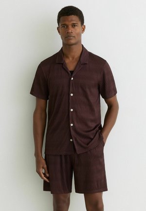 Férfi sötétbarna texturált rövid ujjú gombos ingben és hozzáillő rövidnadrágban, egyik keze a zsebében áll egy fehér fal előtt.