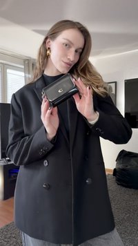 Černá kožená peněženka se zlatým logem, obdélníkový tvar, uzávěr na klopě, držená na pozadí černého saka. Hladká textura, kompaktní design.