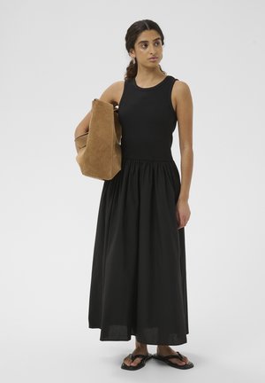 Nő ujjatlan fekete ruhában, kezében egy nagy barna velúr táskával, fekete szandált visel, világos egyszínű háttér előtt áll.