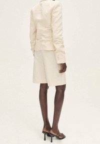 Beige maßgeschneiderter Blazer mit langen Ärmeln, kombiniert mit passenden Shorts. Das Model trägt schwarze Riemchen-Absätze und präsentiert den glatten Stoff und Schnitt.