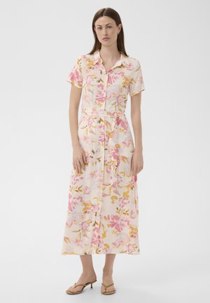 Femme portant une robe midi ceinturée à manches courtes avec col, boutonnée à l'avant, à motif floral rose et jaune, associée à des sandales à talons beiges.