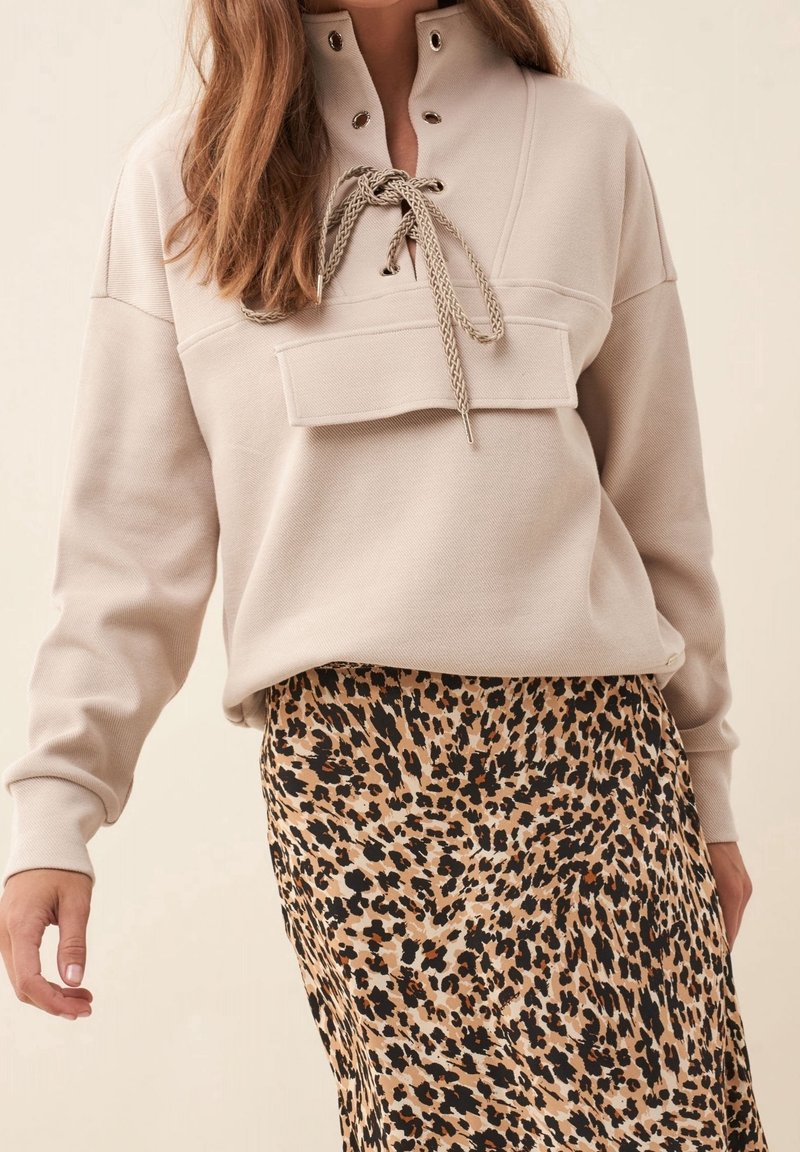 Femme portant un pull beige à col lacé et une jupe à imprimé léopard, debout devant un fond uni.