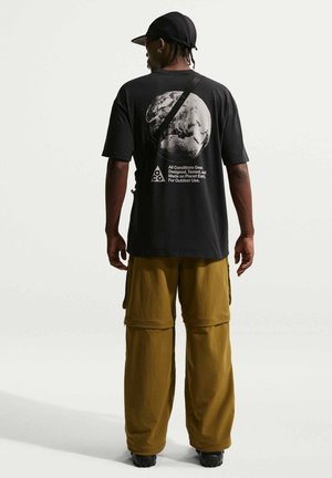 Hombre con gorra negra, camiseta negra con gráfico de la Tierra y texto, pantalones cargo marrones y zapatos negros, visto desde atrás sobre un fondo blanco.