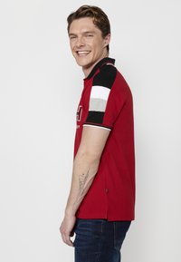 Piros poloing, fekete, fehér és szürke kiemelő ujjal. Gallérral és rövid ujjal rendelkezik. Az elején textúrás logó található. Slim fit fazon.