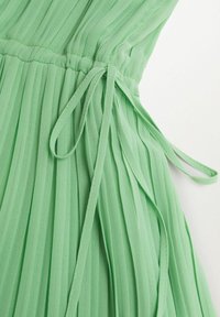 Vestido plisado de tela verde claro, con cintura ceñida y detalle de lazo ajustable, y una textura suave y fluida.