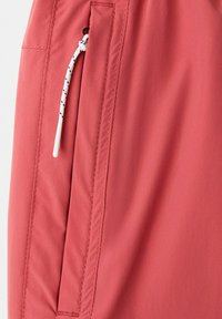 Gros plan sur un pantalon en tissu corail avec une couture verticale et un cordon blanc à pois noirs suspendu près de l'ouverture de la poche.