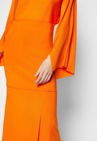 Orange klänning i ett mjukt tyg, med långa klockärmar och en figurnära design med diagonala sömmar och en slits i botten.
