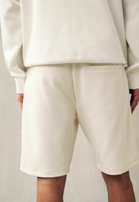 Șorturi de trening de culoare bej deschis, realizate dintr-un material moale, cu un buzunar posterior dreptunghiular și margine rulate. Textură netedă cu detalii minime.