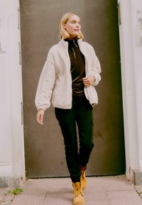 Kreemjas fliisjakis tumeda pruuni särke kohal, koos mustade cargo-pükste ja kollaste saapadega. Sile, pehme tekstuur ja mugav lõige.