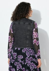 Kamizelka dżinsowa w ciemnoszarym kolorze o dopasowanym kroju. Nałożona na fioletową sukienkę w kwiaty z przezroczystymi rękawami i falbanką na dole.