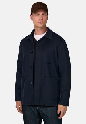 Un homme porte une veste bleu marine boutonnée sur le devant et dotée de trois poches, par-dessus un pull noir et une chemise blanche, avec un jean et une casquette marron.