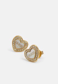 Gouden, hartvormige oorbellen met een witte kern waarop het "GUESS"-logo staat, omringd door een rand van kleine heldere rhinestones.