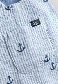 Material albastru cu dungi și suprafață texturată, având broderie cu ancora bleumarin și o etichetă mică „JoJo” pe buzunar.