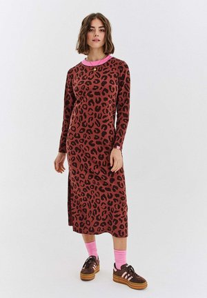 Kobieta ubrana w midi sukienkę w brązowo-czarny leopardi wzór z długim rękawem, różowym kołnierzykiem, różowymi skarpetkami i brązowymi tenisówkami na białym tle.