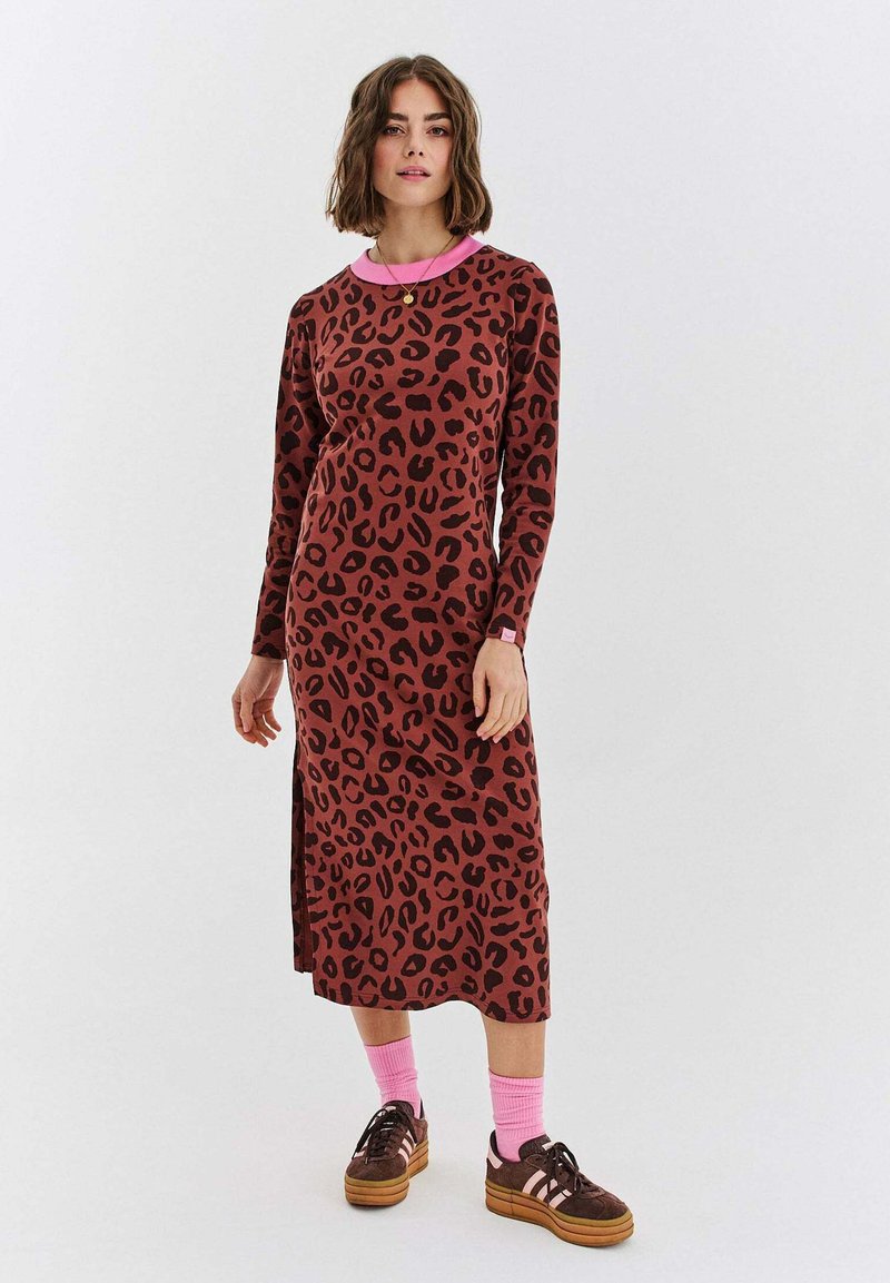 Žena v hnědo-černém leopardím vzorovaném midi šatech s dlouhými rukávy, růžovým límcem, růžovými ponožkami a hnědými teniskami na bílém pozadí.