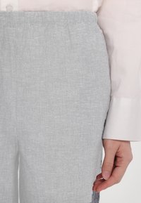 Světlé šedé lněné kalhoty s elastickým páskem; viditelná textura a tkaní látky; spárovány s bílou košilí na knoflíky.