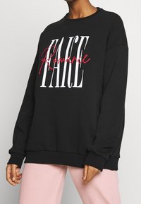 Persona vestida con un sudadera negra que tiene las palabras "Romance" en un rojo cursivo sobre "FAUCE" en letras grandes y blancas, combinada con pantalones rosas.