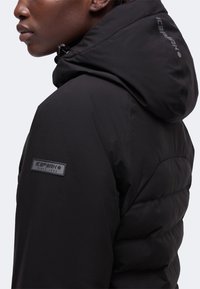 Gros plan d'une personne portant une veste matelassée noire Icepeak avec capuche et écusson de marque sur la manche et la capuche.