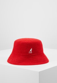Červený klobouk s širokým okrajem vyrobený z texturované tkaniny, s logem Kangol na přední straně. Design má zaoblenou korunku a široký, dolů skloněný okraj.
