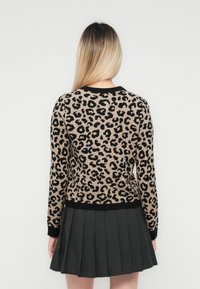 Kvinne med langt blondt hår iført genser med leopardmønster og en svart plisséskjørt, vender bort fra kameraet mot en hvit bakgrunn.