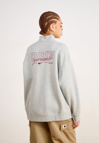 Grå hettegenser med høy krage, med en burgunder "NIKE Sportswear" logo på ryggen. Stoffet er mykt og har en avslappet passform.