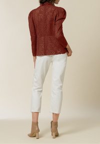 Blusa rossa in pizzo con maniche a palloncino, motivo floreale intricato, abbinata a jeans bianchi alla caviglia e stivaletti color cammello su uno sfondo chiaro.