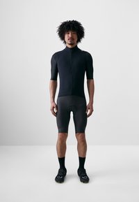 Strój rowerowy z czarnego materiału, z krótkimi rękawami, zapinaną z przodu na zamek i wyściełanymi spodniami. Nosi się go z czarnymi butami i skarpetkami.