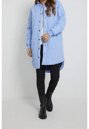 Vrouw die een lichtblauwe gewatteerde jas draagt over een witte top, zwarte strakke broek en zwarte enkellaarzen, staand tegen een effen achtergrond.