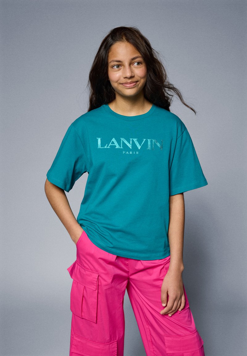 Turquoise oversized t-shirt features "LANVIN PARIS" in silver text. Paired with bright pink cargo pants, showcasing multiple pockets and a relaxed fit.