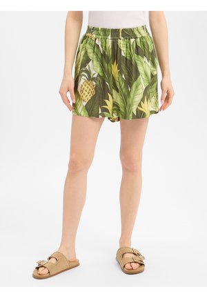 Frau trägt grüne Shorts mit tropischem Blatt- und Ananasmuster, elastischem Taillenbund und beigen Schnallensandalen, steht vor weißem Hintergrund.