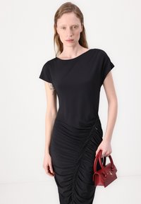 Crna haljina s kapicama, s bočnim naborima i detaljem sa zatvaračem. Model drži malu crvenu torbicu. Glatka tkanina, oblik koji se prilagođava telu.